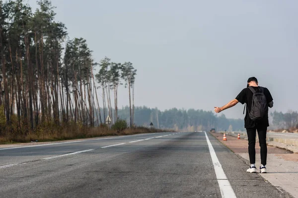 Back view of man with backpack gesturing while hitchhiking on empty road near green trees — Stock Photo