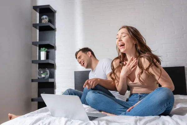 Selective focus of cheerful woman and man laughing while watching comedy movie on laptop in bedroom — Stock Photo
