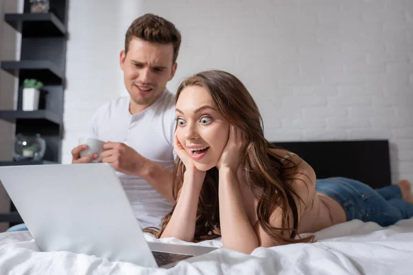 Selective focus of excited woman watching movie on laptop near boyfriend with cup — Stock Photo