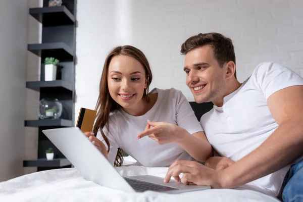 Happy girl pointing with finger at laptop and holding credit card near boyfriend — Stock Photo