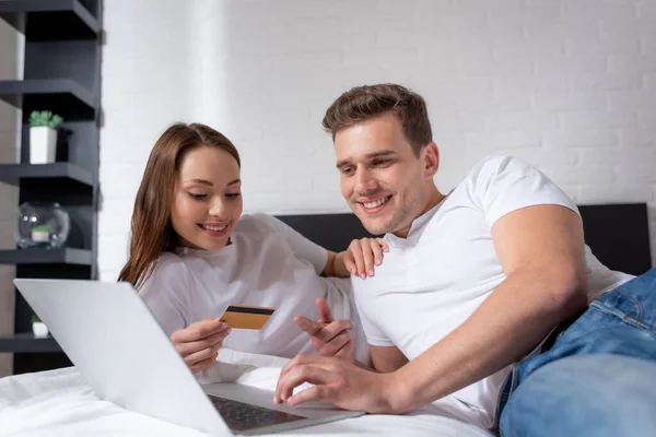 Hombre feliz apuntando con el dedo a la computadora portátil cerca de chica con tarjeta de crédito - foto de stock