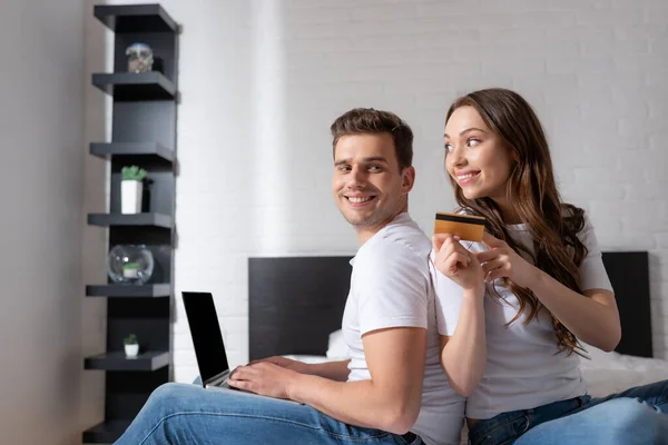 Mujer alegre sosteniendo la tarjeta de crédito y mirando novio usando el teléfono inteligente con la pantalla en blanco - foto de stock