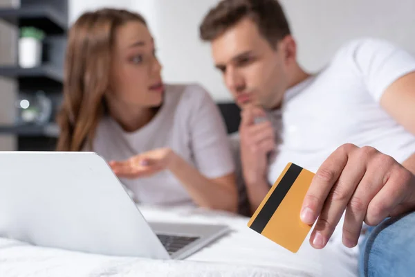 Enfoque selectivo del hombre pensativo que sostiene la tarjeta de crédito cerca de la chica y el ordenador portátil - foto de stock