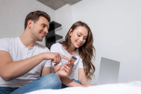Щасливий чоловік вказує пальцем на кредитну картку біля ноутбука і веселої дівчини — стокове фото