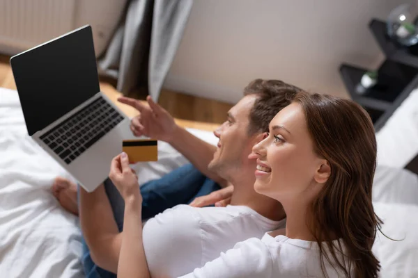 Enfoque selectivo de la mujer alegre que sostiene la tarjeta de crédito cerca del hombre usando el ordenador portátil con la pantalla en blanco en el dormitorio - foto de stock