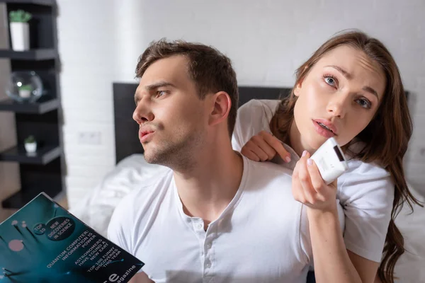 Hombre disgustado sosteniendo la revista mientras se siente caliente cerca de la mujer con controlador remoto de aire acondicionado - foto de stock