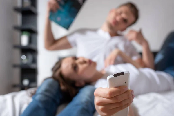 Selective focus of woman with remote controller from air conditioner lying near man holding magazine while feeling hot — Stock Photo