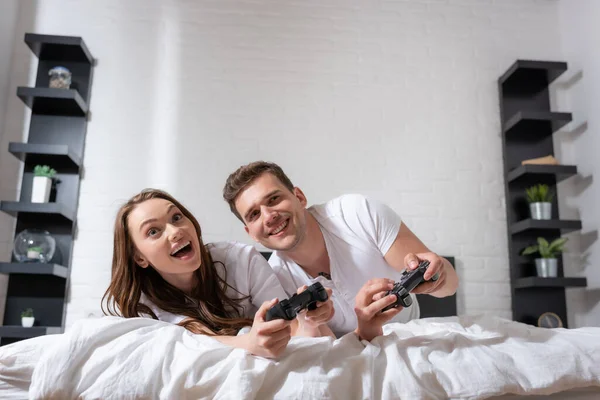 КИЕВ, Украина - 15 мая 2020 года: веселая женщина и счастливый мужчина лежит на кровати и играет в видеоигры — стоковое фото