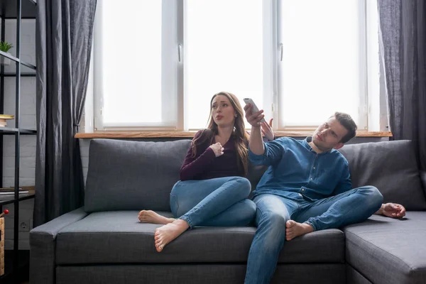 Mulher sentada no sofá perto do homem segurando controlador remoto de ar condicionado enquanto se sente quente — Fotografia de Stock