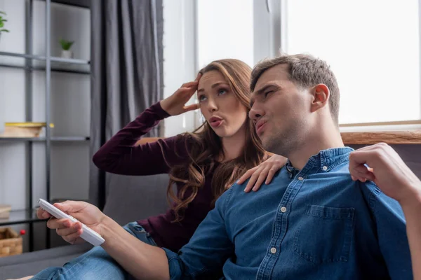 Girl sitting on sofa near displeased man holding remote controller from air conditioner while feeling hot — Stock Photo