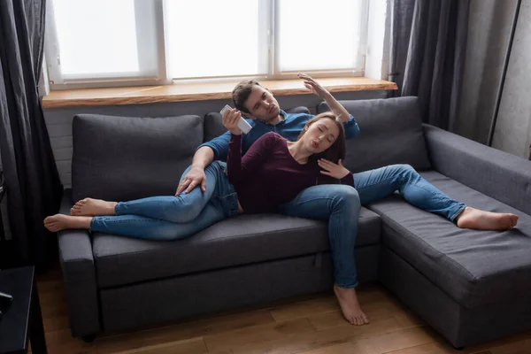 Mujer acostada en el sofá con novio sosteniendo el mando a distancia desde el aire acondicionado mientras se siente caliente - foto de stock