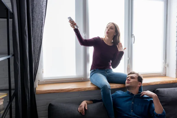 Displeased man near attractive woman holding remote controller from air conditioner while feeling hot — Stock Photo