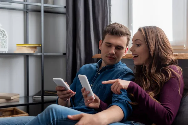 Chica feliz señalando con el dedo en el teléfono inteligente cerca de novio en la sala de estar - foto de stock