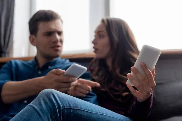 Selective focus of displeased man and beautiful girl holding smartphones in living room — Stock Photo