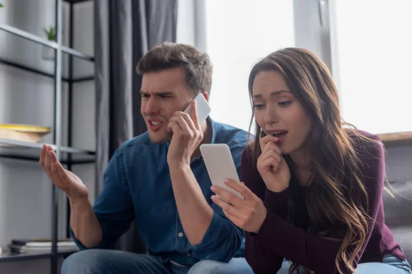 Selektiver Fokus eines überraschten Mädchens, das auf das Smartphone schaut, während ein unzufriedener Mann im Wohnzimmer auf dem Smartphone spricht — Stockfoto