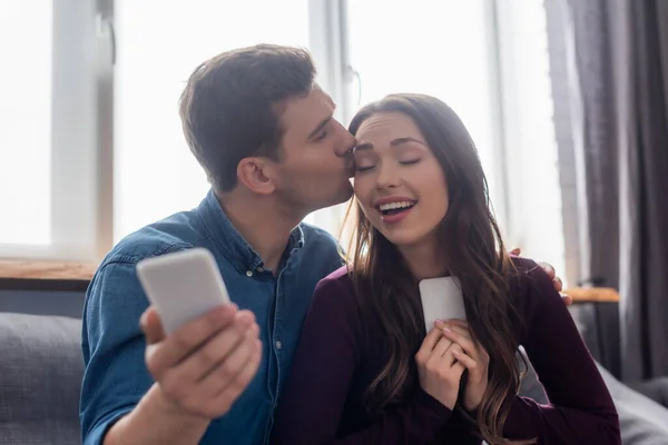 Enfoque selectivo del hombre besando chica feliz y sosteniendo el teléfono inteligente - foto de stock