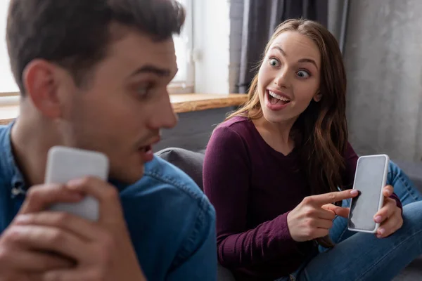 Foyer sélectif de fille excitée pointant du doigt le smartphone avec écran vide et en regardant petit ami dans le salon — Photo de stock