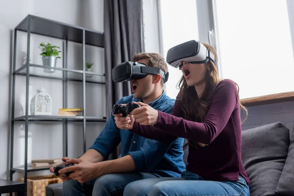 KYIV, UCRANIA - 15 de mayo de 2020: pareja sorprendida con auriculares de realidad virtual que sostienen joysticks mientras juegan videojuegos en la sala de estar - foto de stock