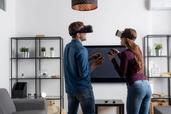 KYIV, UCRANIA - 15 de mayo de 2020: pareja alegre en auriculares de realidad virtual con joysticks y jugando videojuegos en la sala de estar - foto de stock