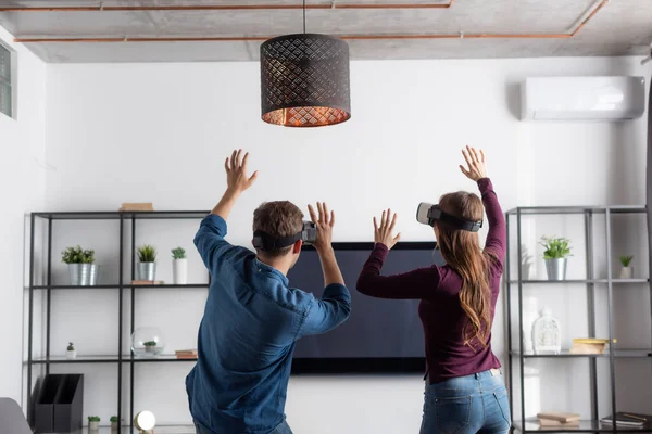 Vista trasera de la pareja en auriculares de realidad virtual haciendo gestos mientras se juega en la sala de estar - foto de stock