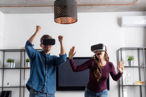 Happy couple in virtual reality headsets celebrating triumph while gaming in living room — Stock Photo