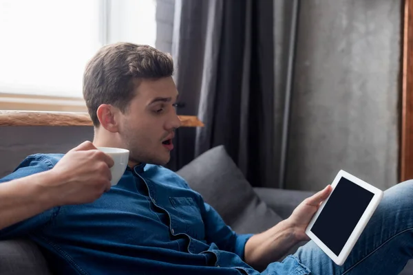 Hombre sorprendido sosteniendo taza y mirando tableta digital con pantalla en blanco - foto de stock