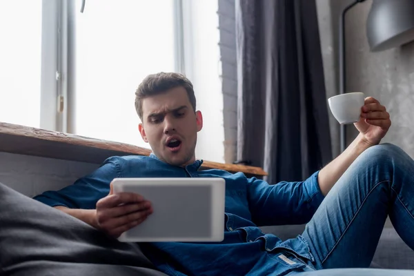 Вибірковий фокус шокованого чоловіка тримає чашку кави і дивиться на цифровий планшет — стокове фото