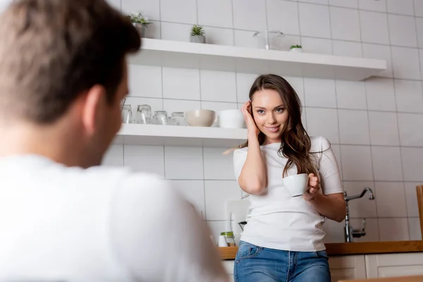 Messa a fuoco selettiva di ragazza toccando i capelli e guardando l'uomo mentre tiene la tazza — Foto stock