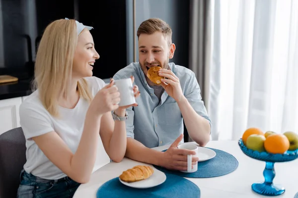 Happy girl holding cup and looking at boyfriend eating croissant — Stock Photo