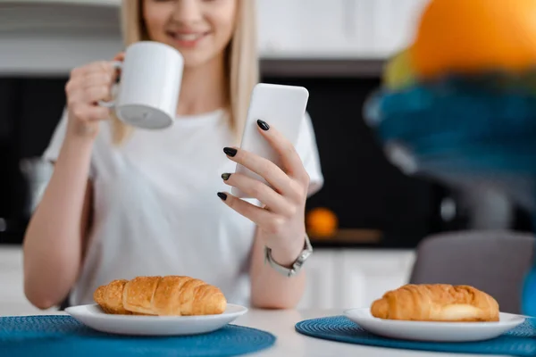 Vista recortada de niña sosteniendo teléfono inteligente y taza cerca de croissants - foto de stock