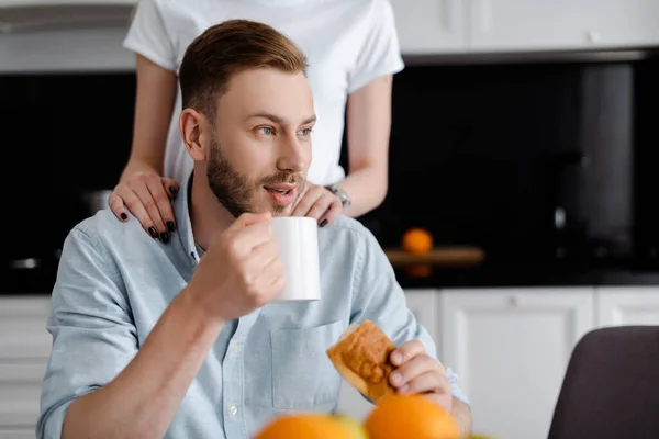 Enfoque selectivo de chica tocando novio guapo sosteniendo taza de café y croissant - foto de stock