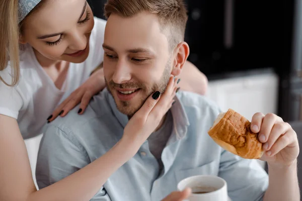 Selective focus of beautiful girl touching handsome boyfriend holding cup of coffee and croissant in kitchen — Stock Photo