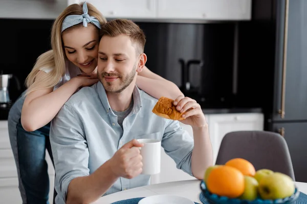 Selektiver Fokus der schönen Mädchen, die fröhlichen Freund mit einer Tasse Kaffee und Croissant in der Küche berühren — Stockfoto