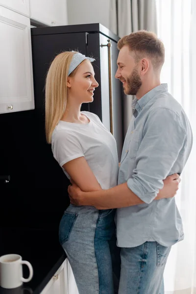 Happy girlfriend and bearded boyfriend looking at each other while hugging in kitchen — Stock Photo