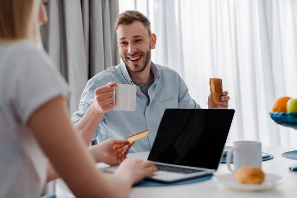 Enfoque selectivo de la niña sosteniendo la tarjeta de crédito cerca de la computadora portátil con pantalla en blanco y novio feliz - foto de stock