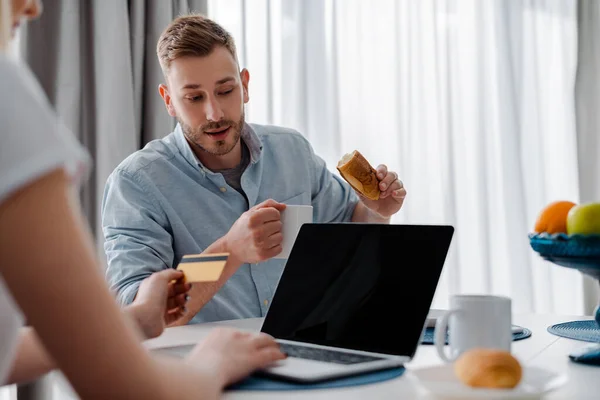 Enfoque selectivo de la mujer que sostiene la tarjeta de crédito cerca del ordenador portátil con pantalla en blanco y novio guapo con taza y pastelería - foto de stock