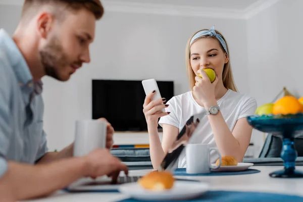 Enfoque selectivo de la niña comiendo manzana y el uso de teléfonos inteligentes cerca freelancer novio y desayuno - foto de stock
