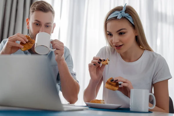 Selektiver Fokus attraktiver Mädchen mit Croissant in der Nähe von Freelancer-Freund, der Kaffee in der Nähe von Laptop trinkt — Stockfoto