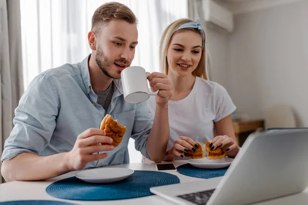 Foyer sélectif de joyeux couple de pigistes prenant le petit déjeuner et regardant ordinateur portable — Photo de stock