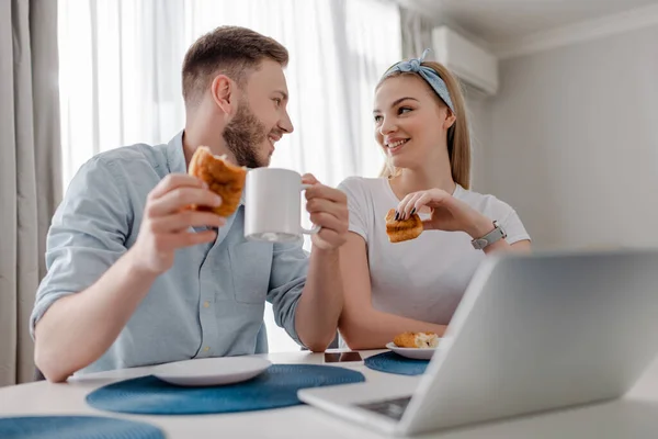 Enfoque selectivo de alegre pareja de freelancers desayunando y mirándose el uno al otro cerca del portátil - foto de stock
