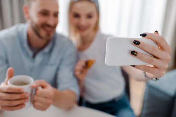 Enfoque selectivo de la mujer feliz sosteniendo el teléfono inteligente y tomar selfie con el novio - foto de stock