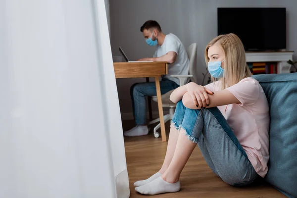 Foyer sélectif de la fille dans le masque médical assis sur le sol près de copain pigiste travaillant à la maison — Photo de stock