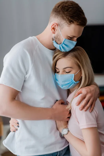 Couple dans des masques médicaux tenant la main et étreignant à la maison — Photo de stock