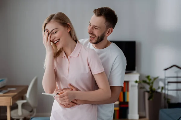 Счастливый мужчина обнимает веселую девушку проведение теста на беременность с положительным результатом — стоковое фото