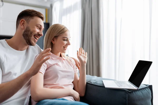 Alegre pareja saludando las manos y mirando a la computadora portátil con la pantalla en blanco, mientras que tener chat de vídeo - foto de stock