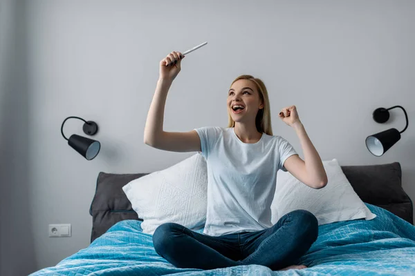 Mujer excitada sosteniendo la prueba de embarazo con resultado positivo y sentada en la cama - foto de stock