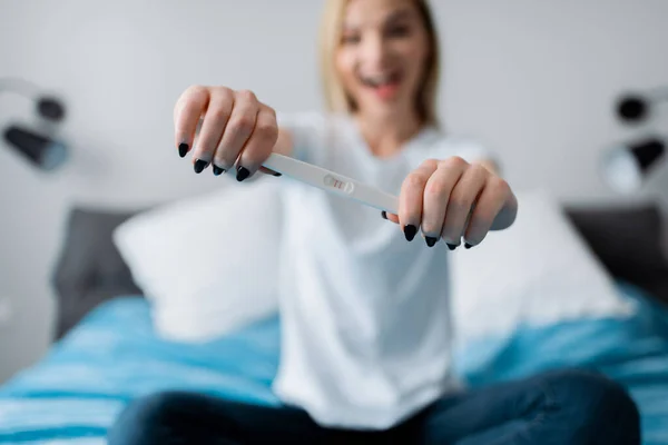 Selective focus of excited woman holding pregnancy test with positive result and sitting on bed — Stock Photo