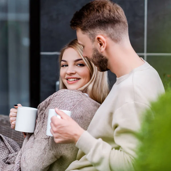 Selektiver Fokus des glücklichen Mädchens, das eine Tasse in der Hand hält und in die Kamera neben dem bärtigen Freund blickt — Stockfoto