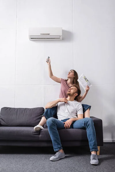 Casal exausto com controlador remoto e jornal sofrendo de calor enquanto sentado em casa com ar condicionado quebrado — Fotografia de Stock