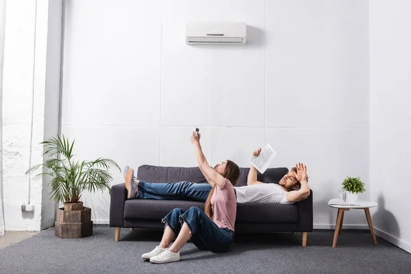 Pareja joven con mando a distancia y periódico que sufre de calor mientras se sienta en casa con el aire acondicionado roto - foto de stock